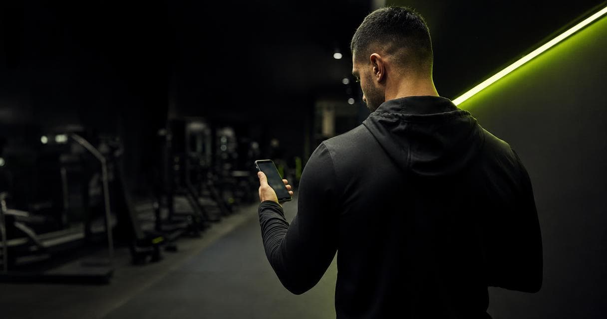 Online fitness coach comparing HubFit alternatives on his phone in a dark premium gym with a lime-green LED strip